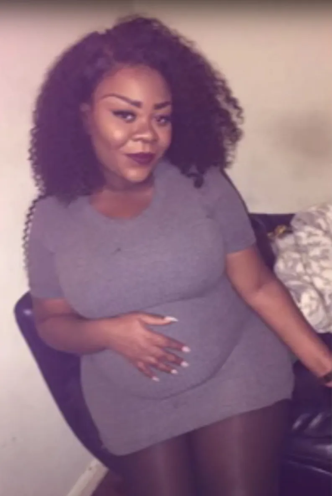 A woman with dark curly hair, wearing a gray t-shirt dress and dark tights, sits with her hand on her stomach.