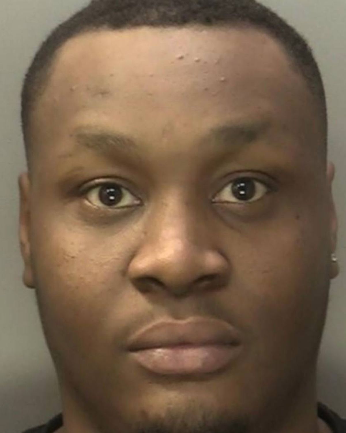 An image depicts a close-up view of a person's face, focusing on the upper half. The individual displays a serious expression, with clearly visible eyes, a straight nose, and defined lips. The skin tone is brown, and the background is plain, indicating this may be a mugshot or a similar official photograph.