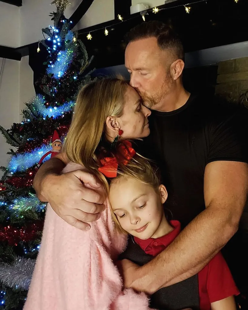 Mel Schilling embraces her family, a man kissing her forehead and a young girl leaning on her, in front of a Christmas tree.
