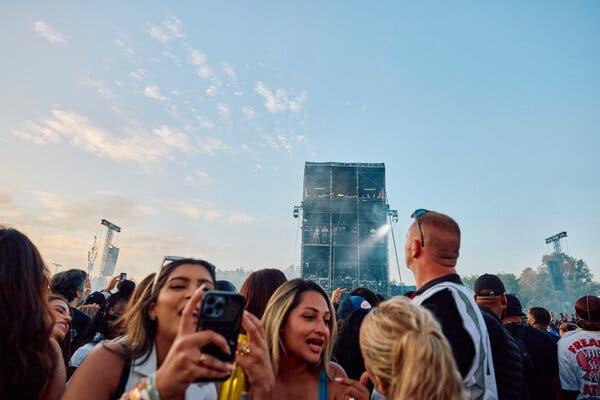 People stand in a crowd in an open area in daylight, some holding up their phones to take photos.