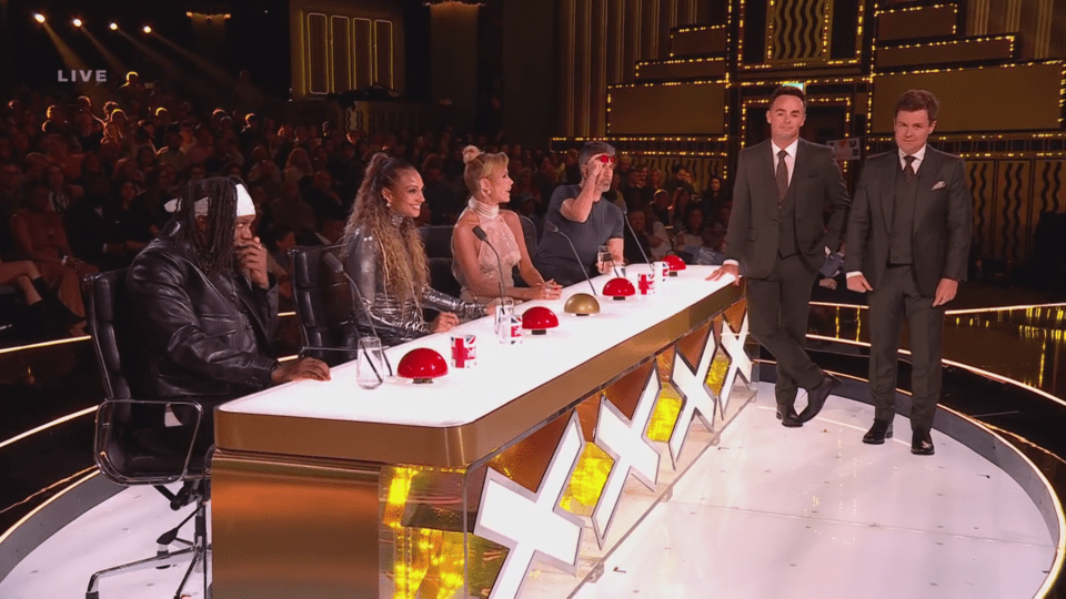 Two men in suits standing beside a table where four judges are seated with buzzers.