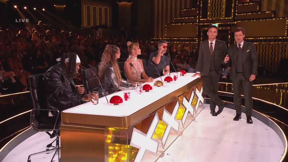 Four judges and two hosts stand on a brightly lit stage with an audience in the background.