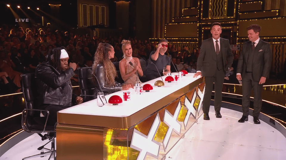 A panel of four judges and two hosts stand on a stage with a live audience in the background.