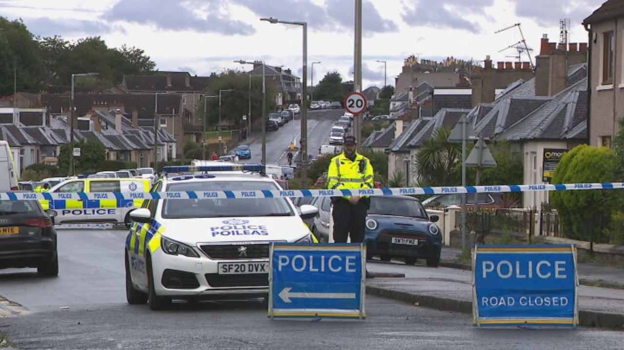 A residential street in Edinburgh cordoned off with police tape. There are blue signs that read police road closed and several police cars and vans behind the tape while one officer stands guard.