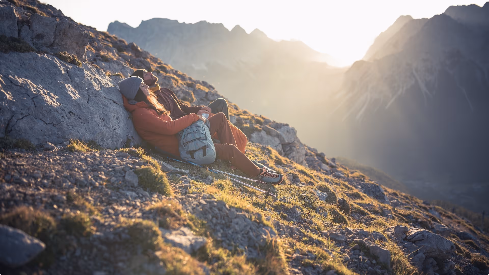Zwei Wanderer sitzen entspannt auf einem Berghang, genießen den Sonnenuntergang in den Alpen, Berglandschaft im Hintergrund.