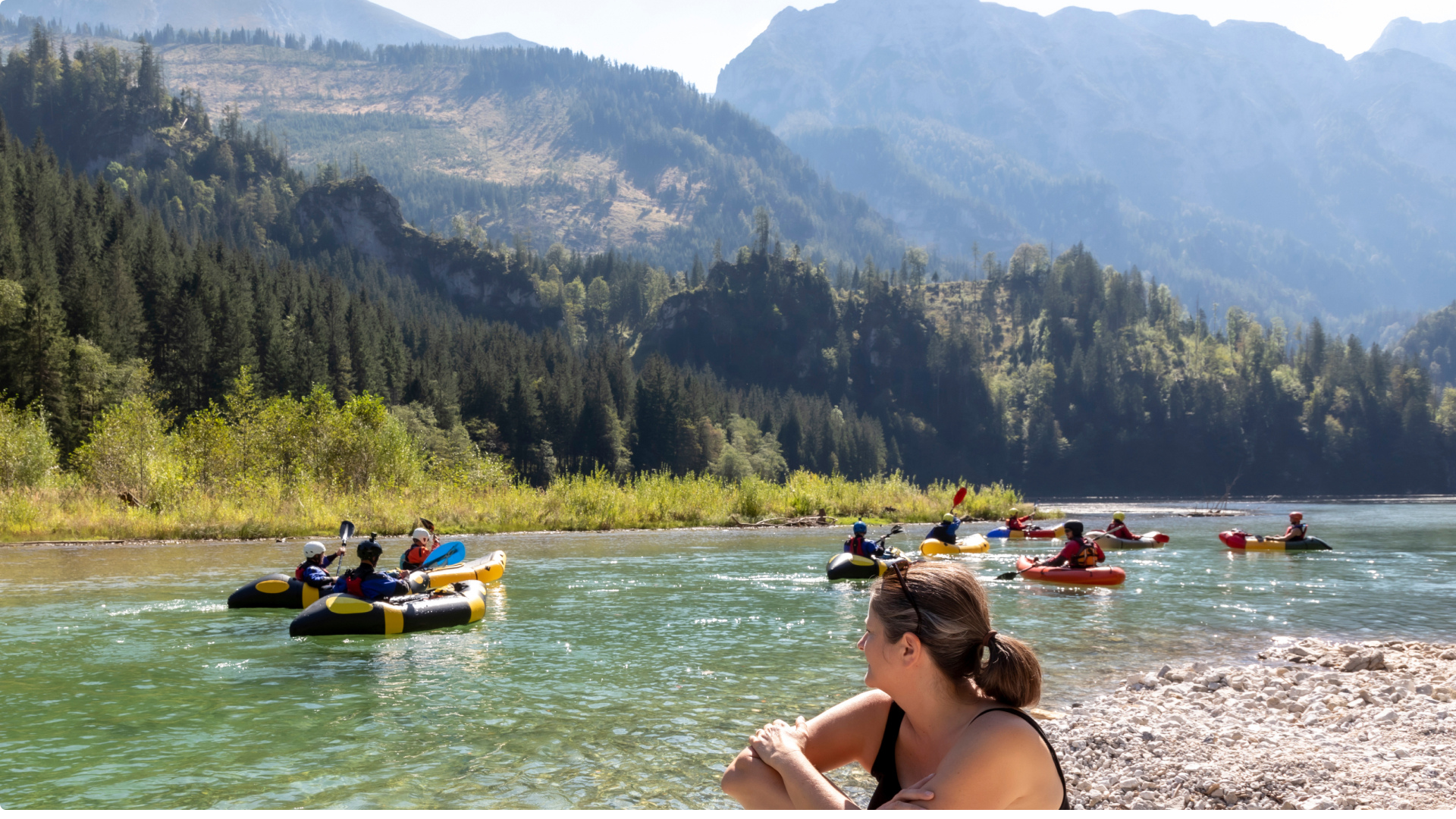 Frau sitzt an der Mur und schaut den Raftingbooten zu