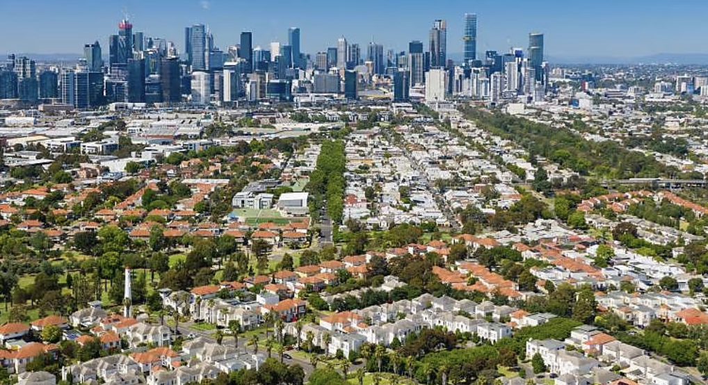 Aerial view of Melbourne city skyline with residential neighbourhoods, tree-lined streets, and modern skyscrapers under a clear blue sky.