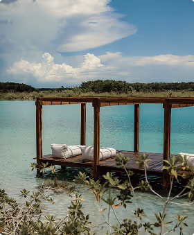 Wooden dock with white cushions extending over turquoise water under a partly cloudy sky.