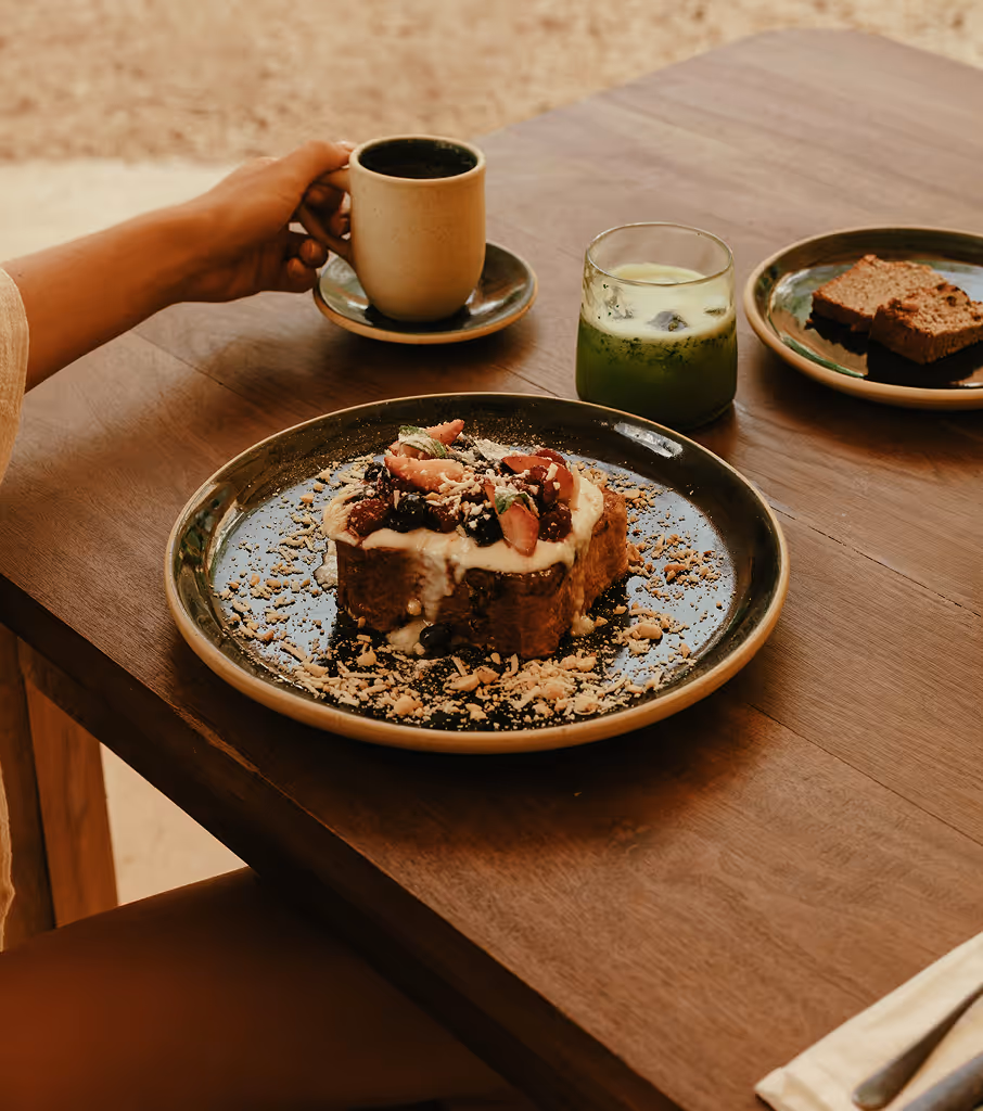 Plate of French toast topped with cream, berries, and mint, alongside a cup of coffee, a glass of green drink, and two pieces of cake on a wooden table.