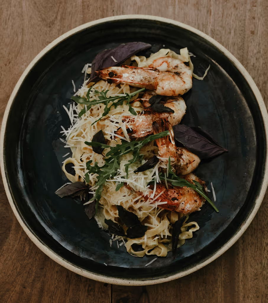 Black plate with pasta topped with grilled shrimp, grated cheese, and mixed greens on a wooden table.