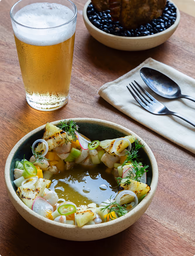 A bowl of fresh ceviche with grilled pineapple, sliced onions, jalapeños, and herbs on a wooden table next to a glass of beer and cutlery on a napkin.