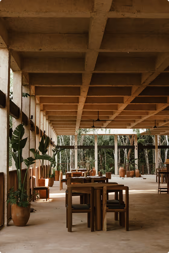 Open-air dining area with wooden tables and chairs, large potted plants, and a concrete ceiling with beams.