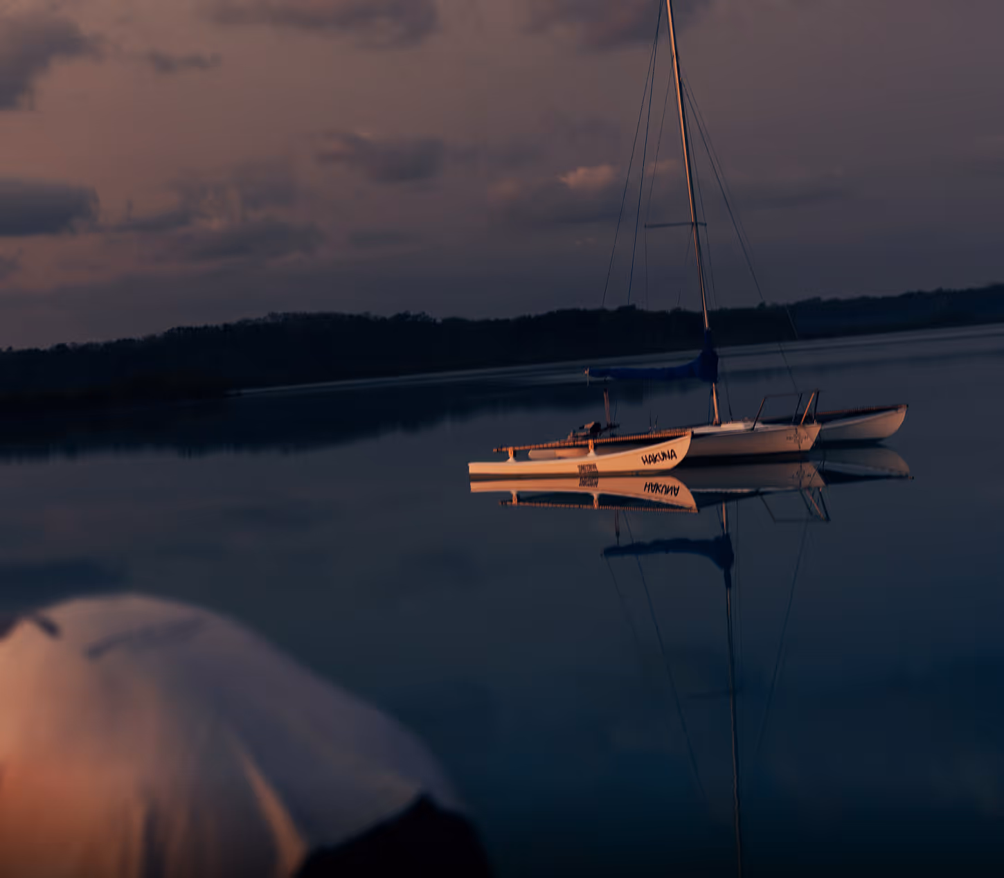 Sailboat named Hakuna floating on calm water at dusk with a reflection of the boat and mast.
