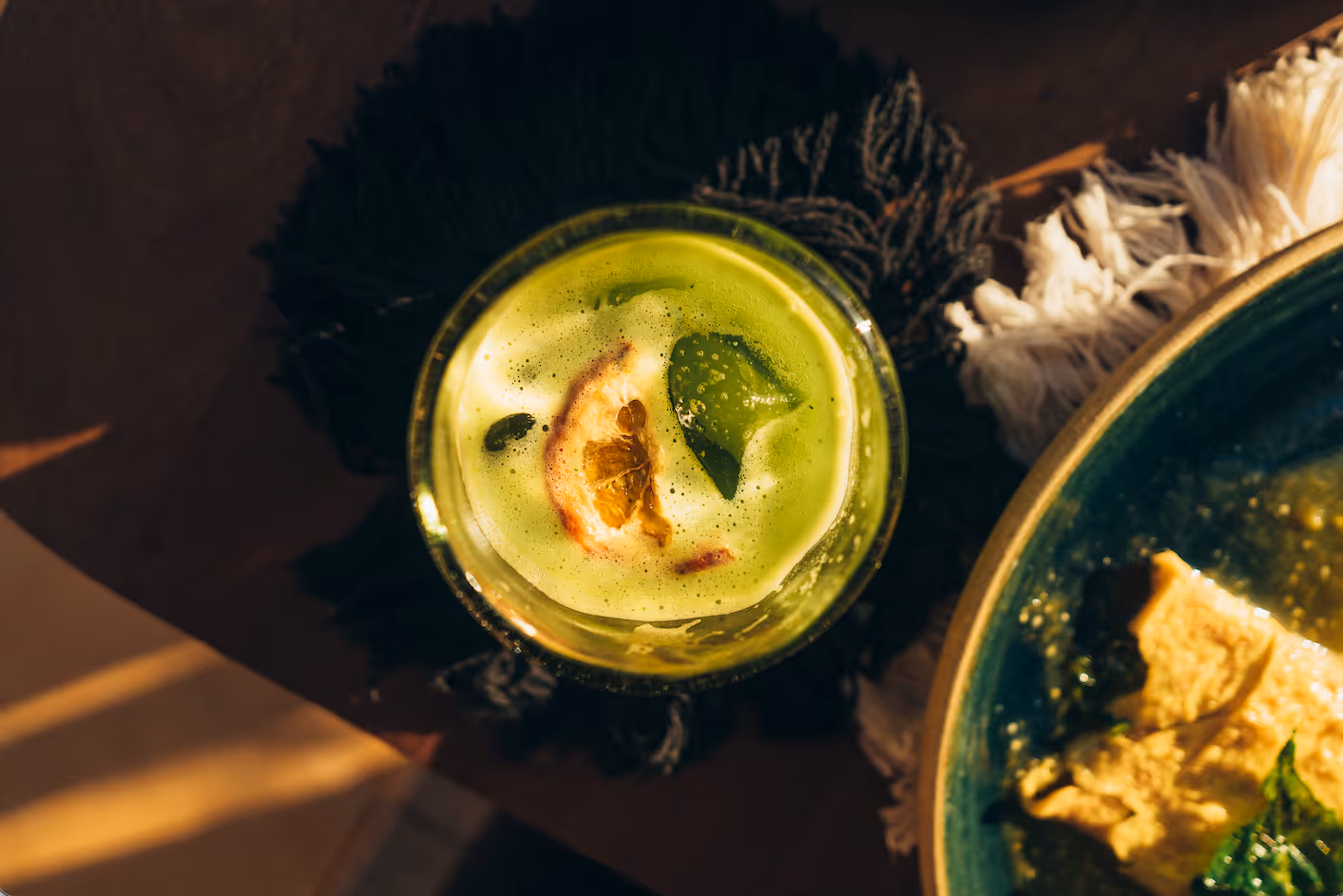 Top view of a frothy green matcha drink with a lemon slice and mint leaf in a glass next to a bowl with food.
