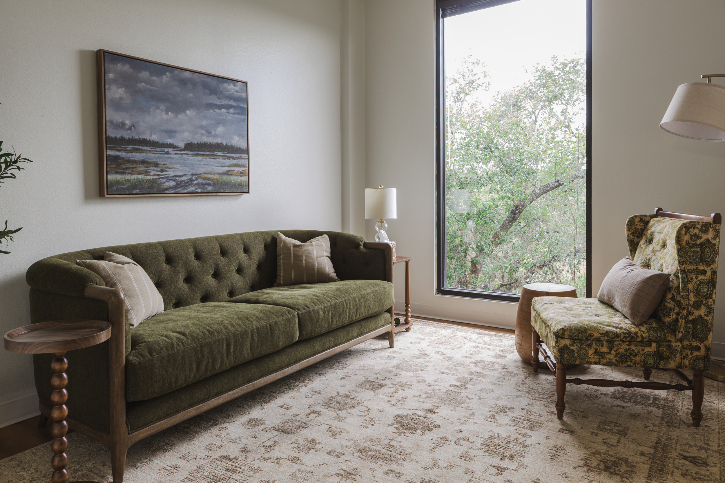 Counseling office with olive green tufted sofa, floral wingback chair, landscape painting, and tall window overlooking trees