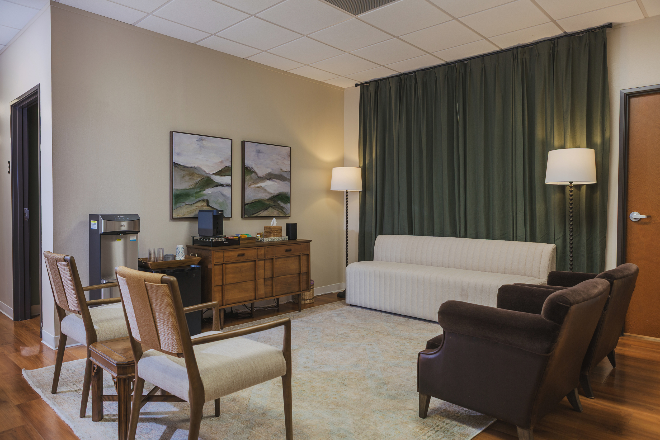 Therapy room with cream sofa, dark brown armchairs, wooden chairs, large patterned rug, and dark green curtains