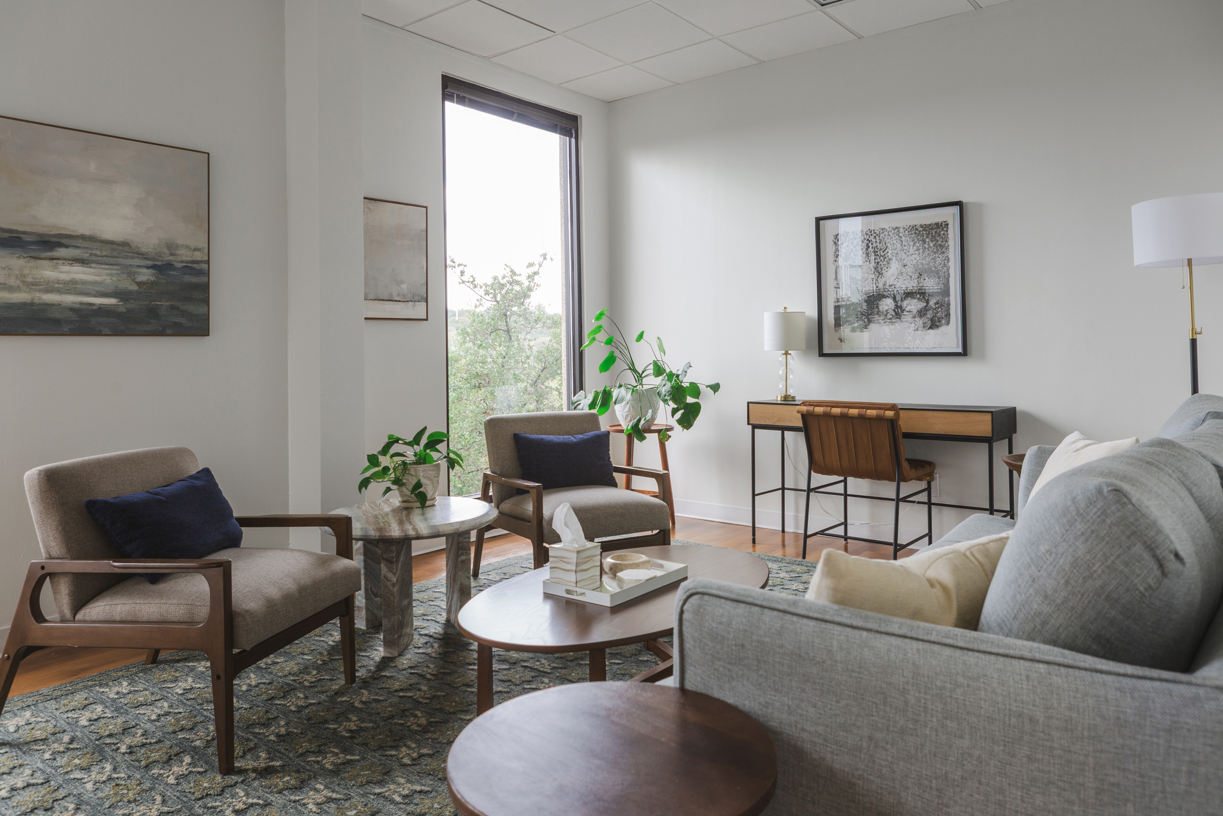 Therapy office with two tan armchairs, navy pillows, oval wood coffee table, patterned rug, and window to green trees