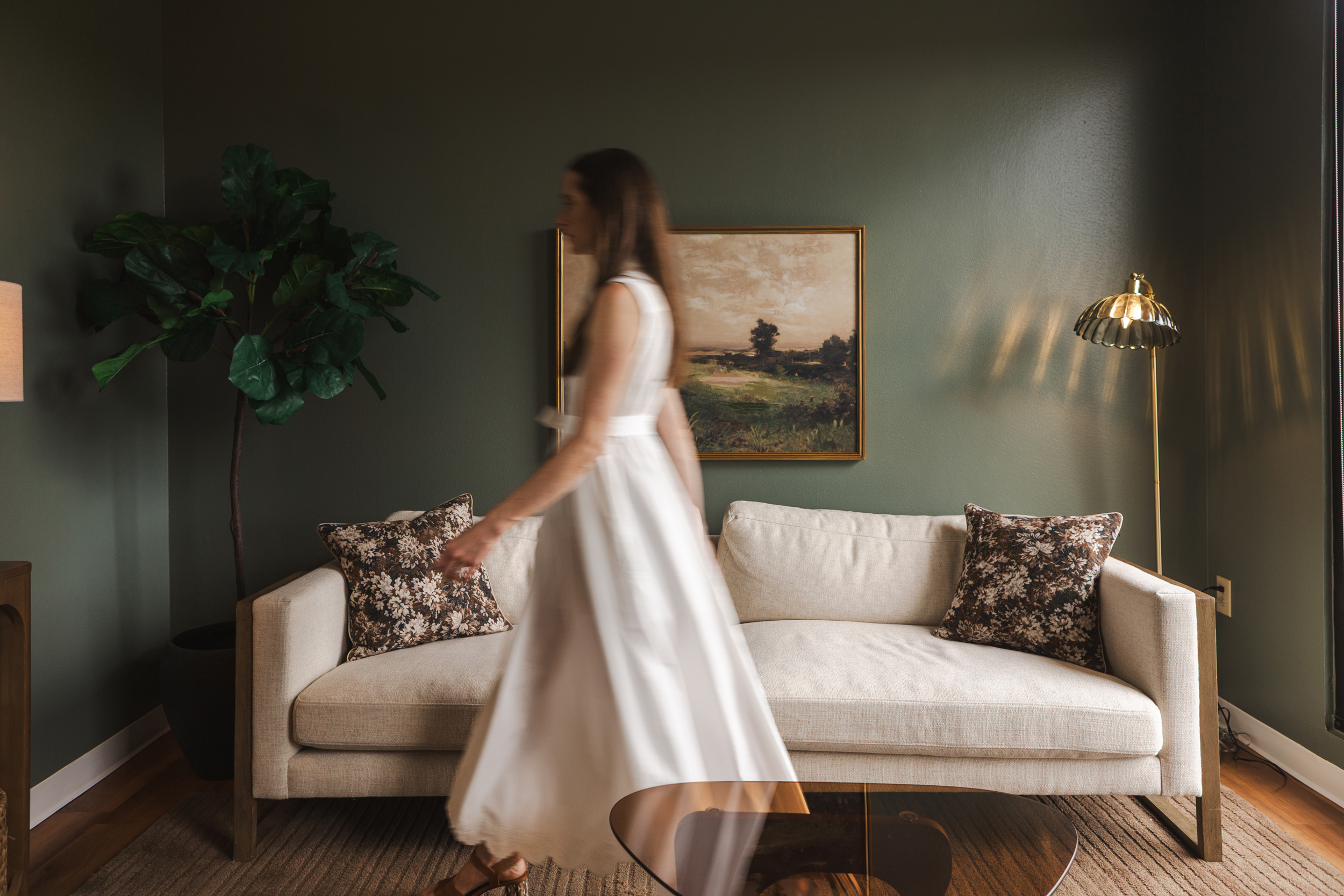 Therapy office with cream sofa against olive green wall, patterned pillows, landscape painting, and gold floor lamp
