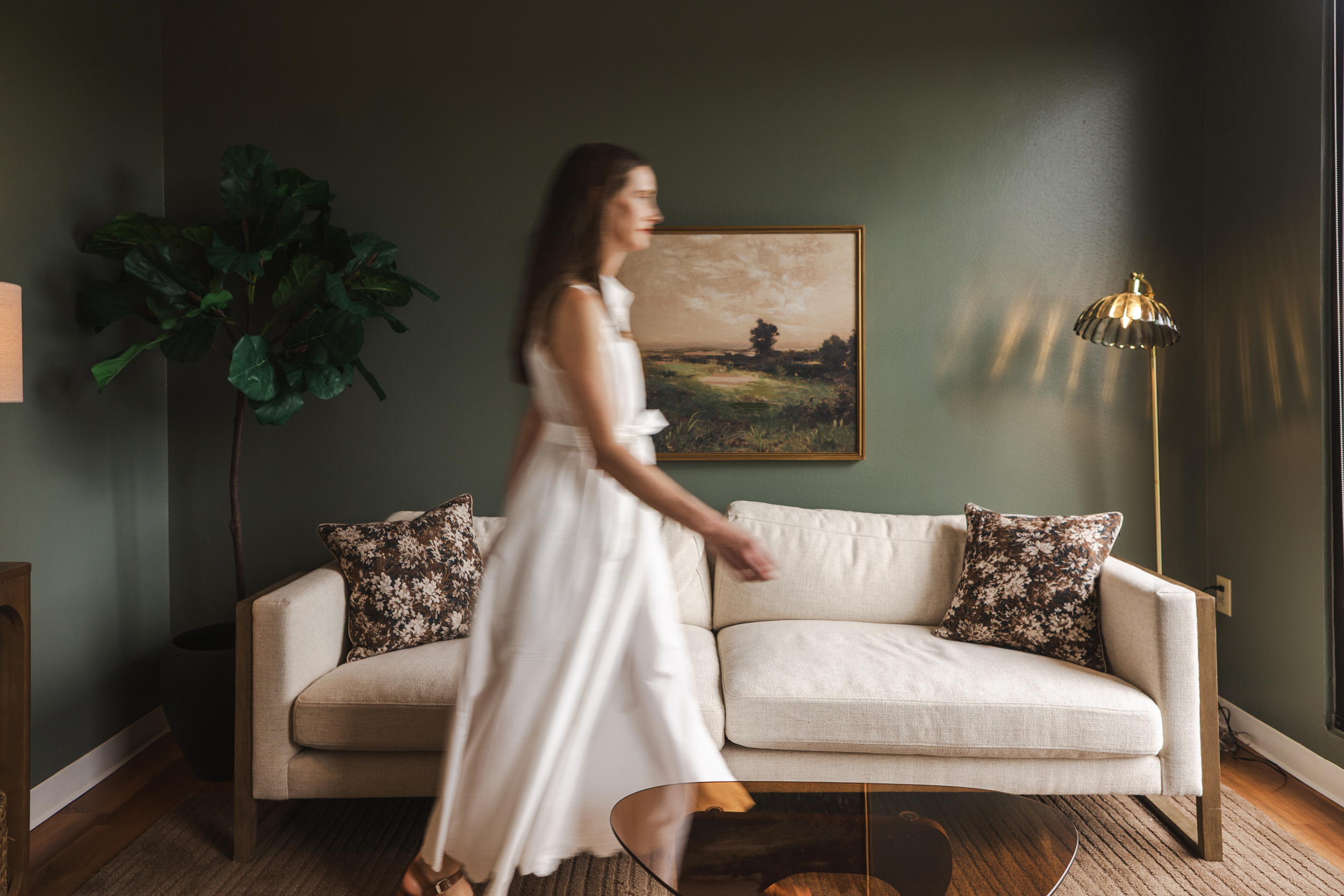 Office interior with white wood-frame sofa, botanical pillows, landscape art, and sage green walls