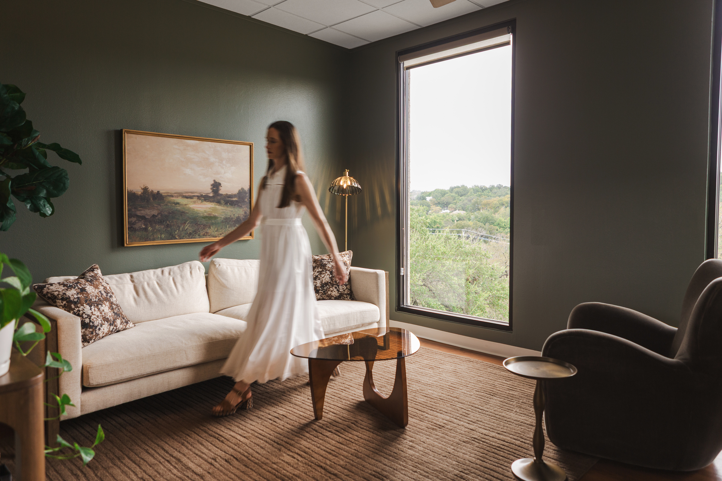 Therapy room with olive green walls, cream sofa, mid-century wood coffee table, and window overlooking trees