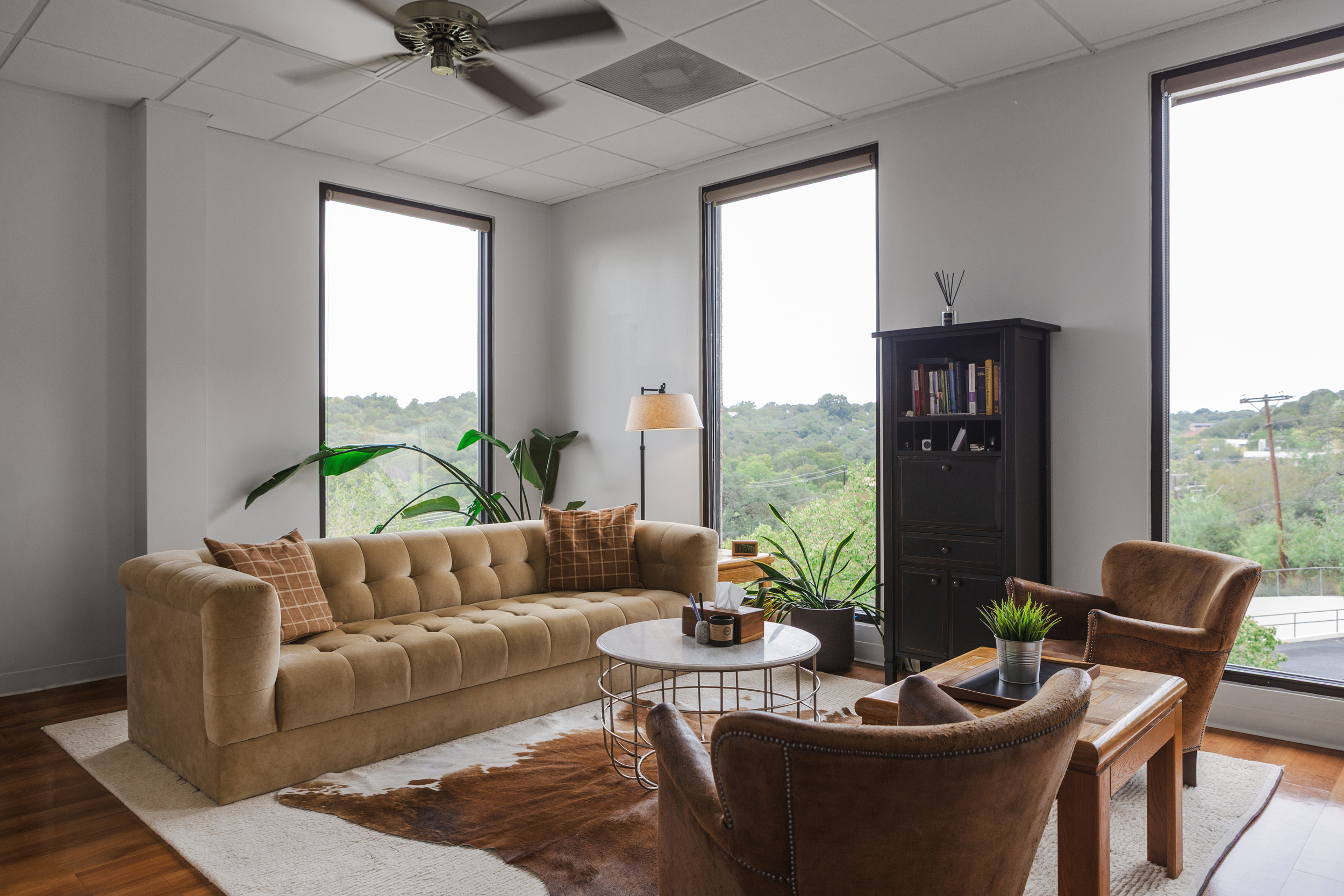 Modern counseling office with mustard velvet sofa, brown armchairs, marble coffee table, and windows to green hills