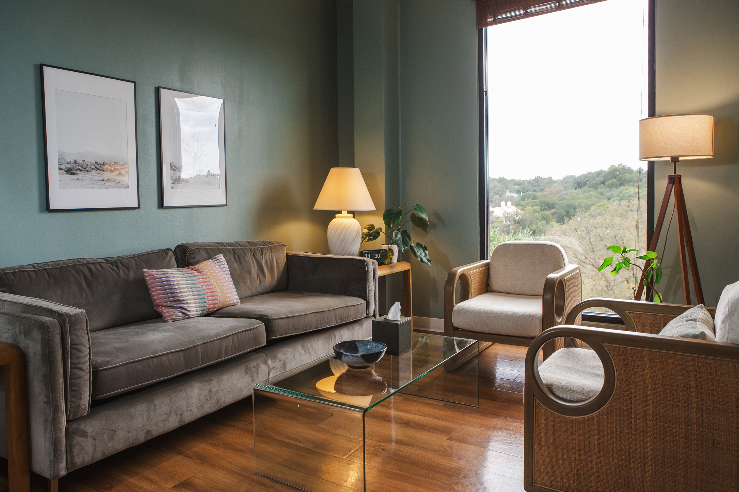 Therapy office with gray velvet sofa, wooden armchairs with cream cushions, glass coffee table, and sage green walls