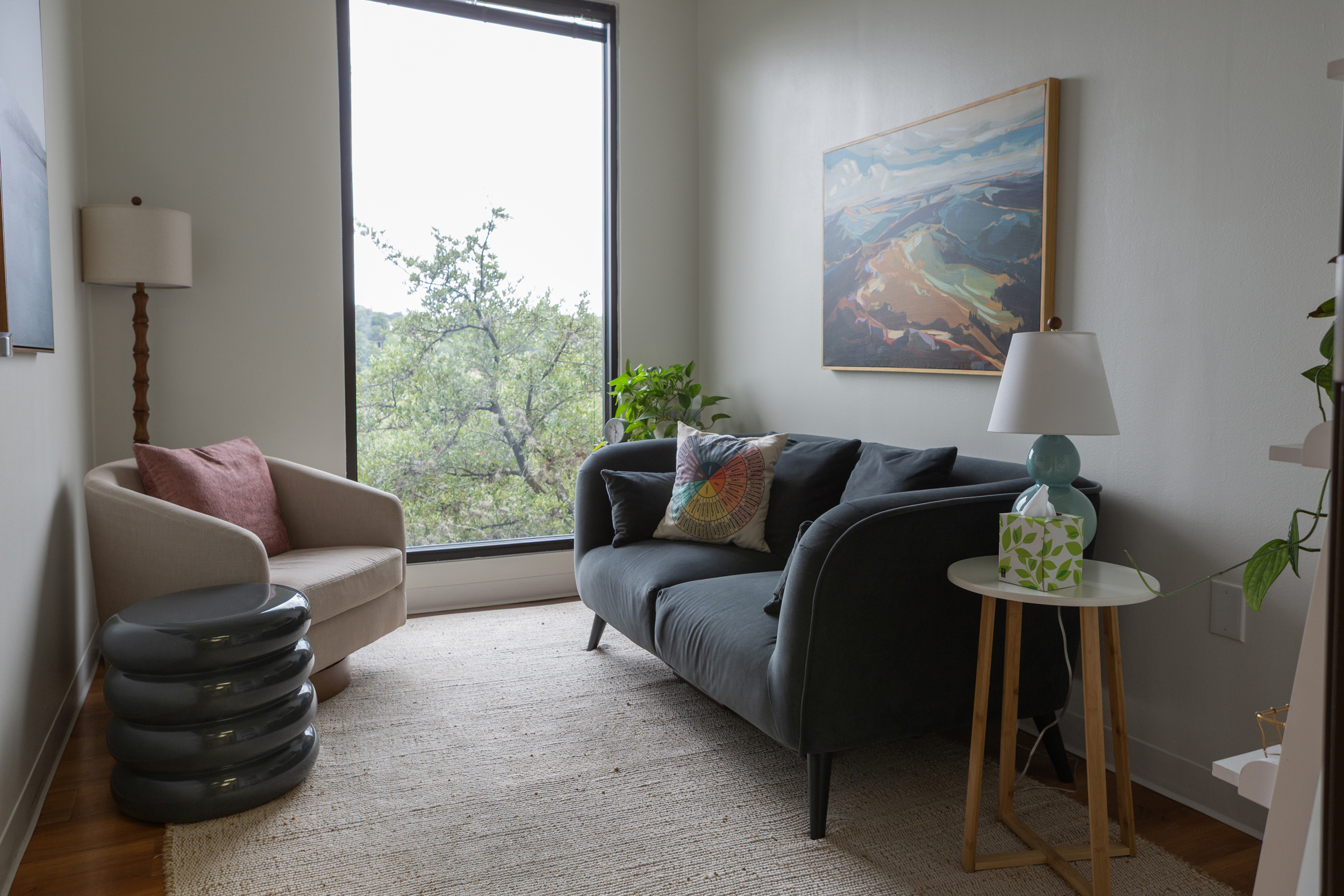 Bright therapy room with dark gray loveseat, tan armchair, landscape painting, and large window to green trees
