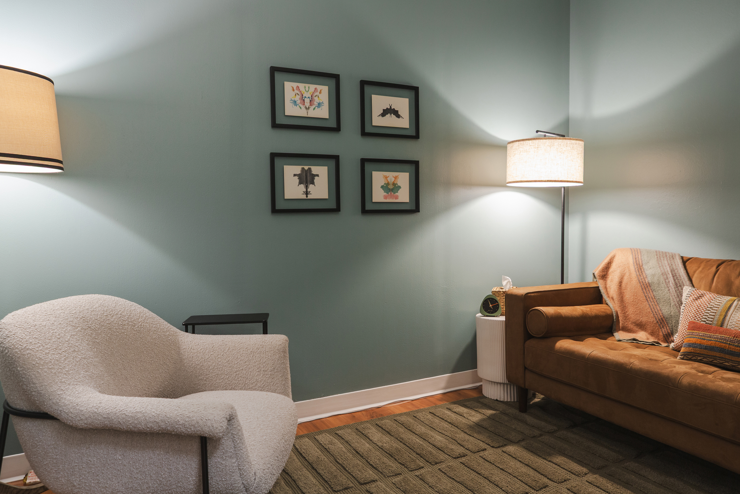 Cozy therapy room with white boucle armchair, brown leather sofa, teal walls, and framed art grid