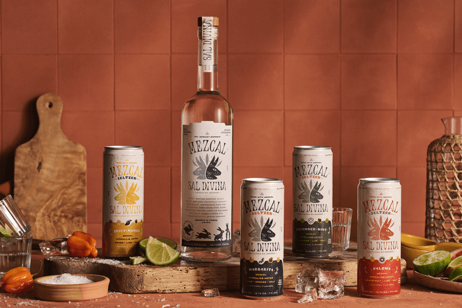 Four cans of Sal Divina's Mezcal seltzers in various flavors and a tall bottle of Mezcal in a composition with cutting boards, ice, salt, limes, and peppers.