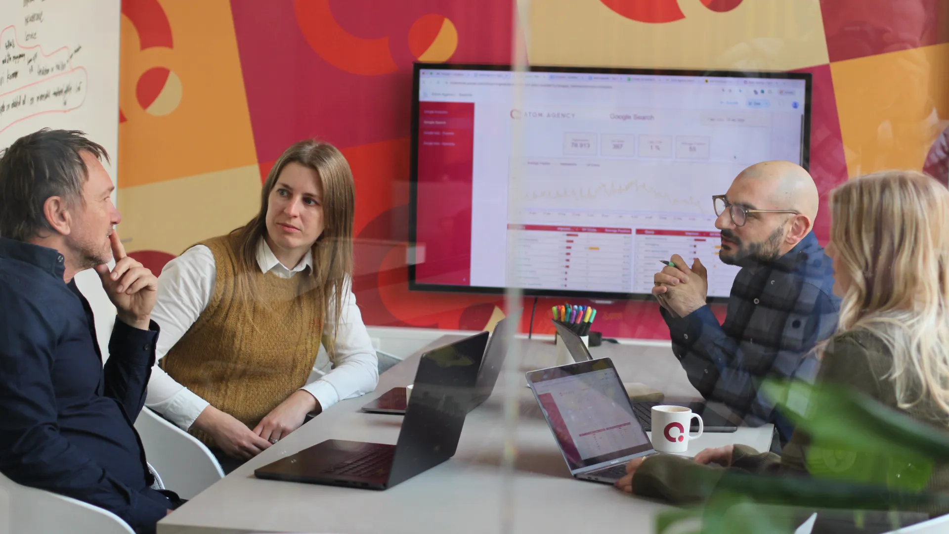 Människor i ett möte med bärbara datorer och en stor skärm som visar data och grafer på väggen.