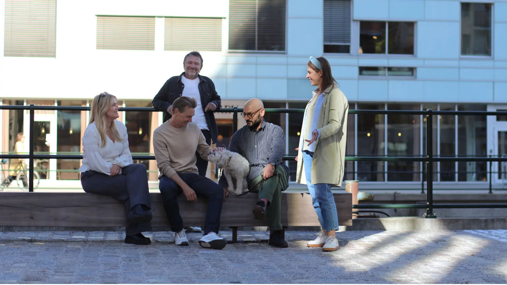 Människor som sitter och står utomhus med en hund på en solig dag i stadsmiljö.