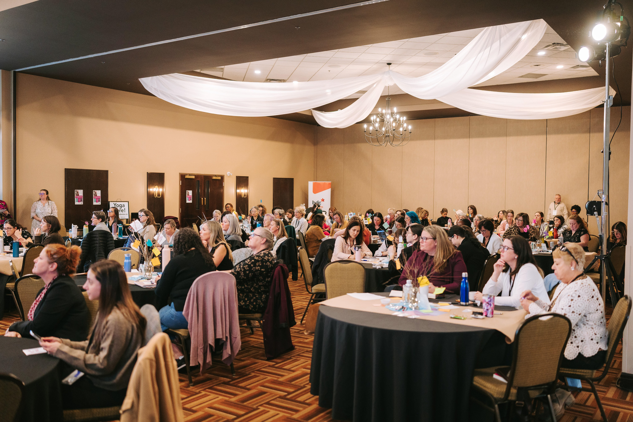 Large group of people seated at round tables in a conference room with beige walls and draped ceiling fabric.