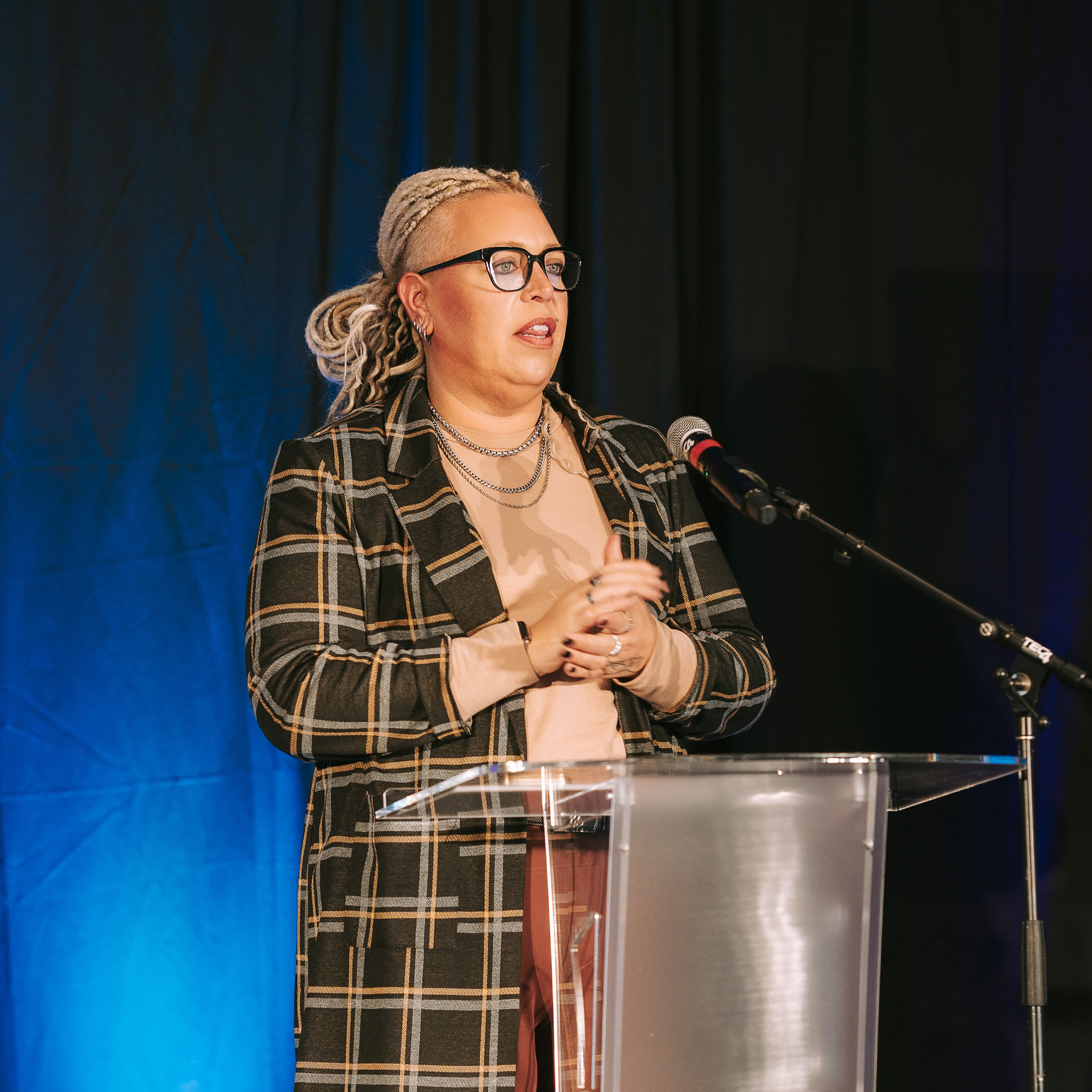Chris McMartin speaking at a clear podium with a microphone, against a dark background with blue lighting.