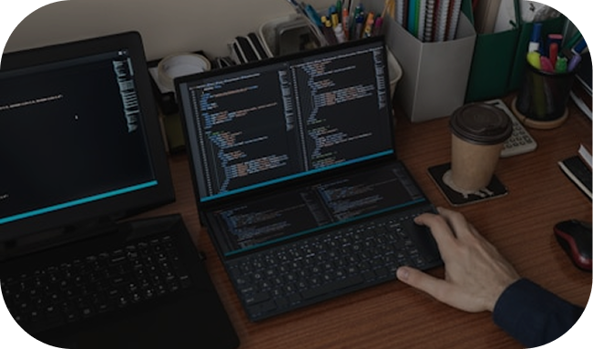 Person typing on a dual-screen laptop with programming code displayed on both screens, next to a coffee cup on a wooden desk.