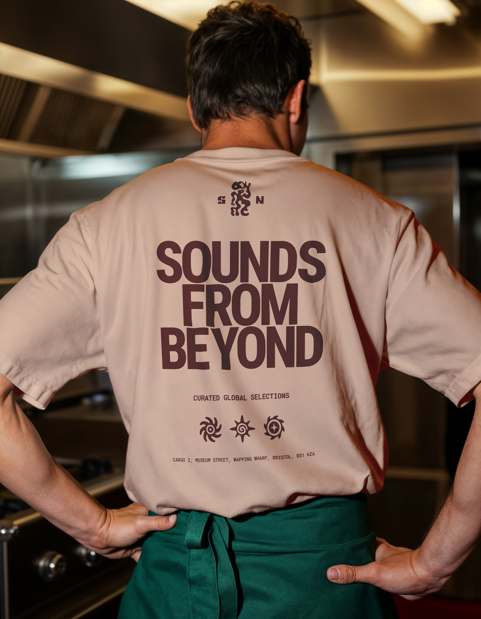 Person wearing a light pink t-shirt with large dark text reading 'SOUNDS FROM BEYOND' and smaller text and symbols beneath, paired with a green apron in a kitchen setting.