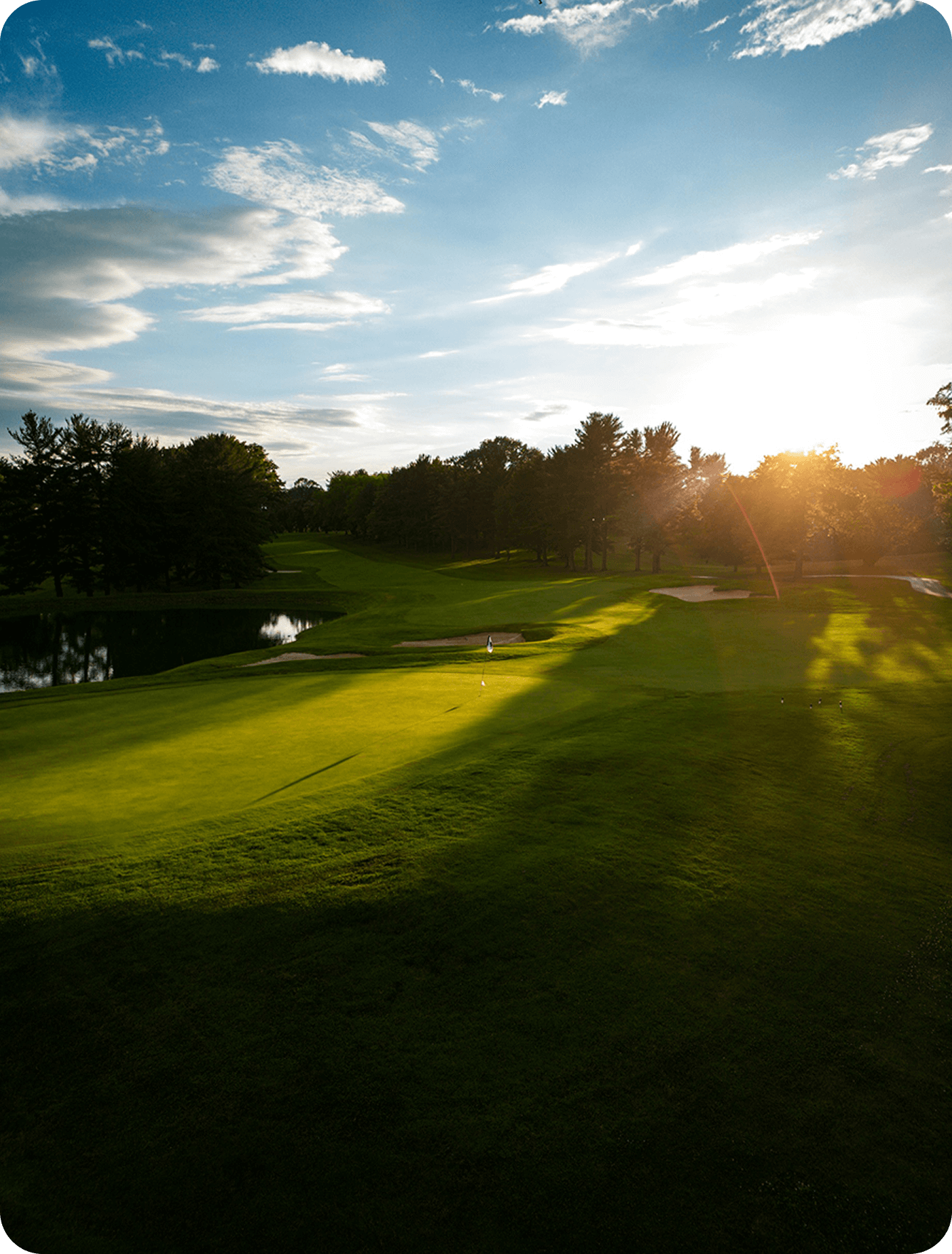 Sunset over a lush green golf course with a putting green, sand bunkers, trees, and a small water hazard.