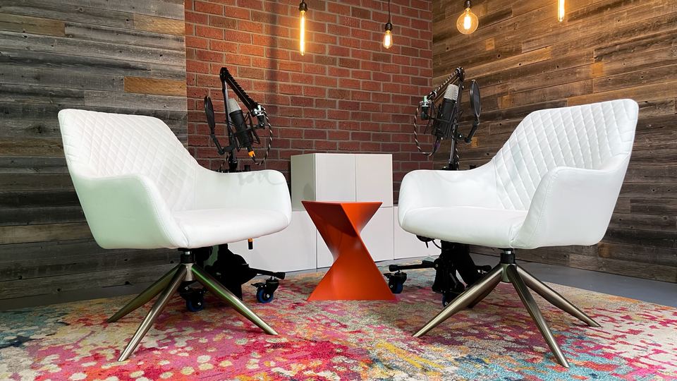 Interview set in Studio A with two white armchairs in front of a brick and wood wall with hanging lamps.