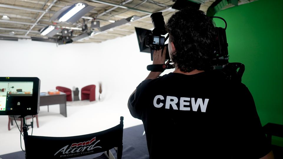 Camera operator seen from behind filming on the Studio A office set, next to the green screen.