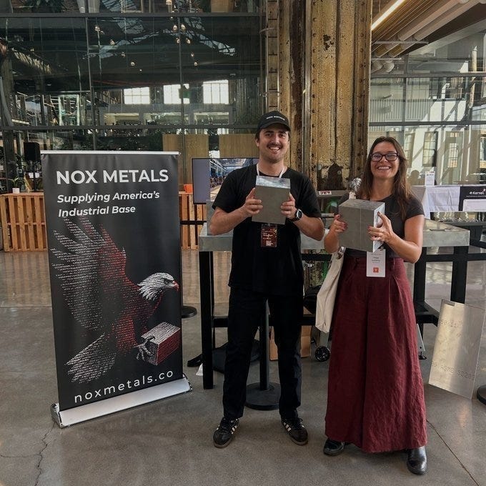 Two people standing in an industrial setting, holding metal blocks. A banner displays "Nox Metals" with text "Supplying America\'s Industrial Base" and an eagle graphic. The background includes large windows, metal beams, and a check-in desk with additional signage.