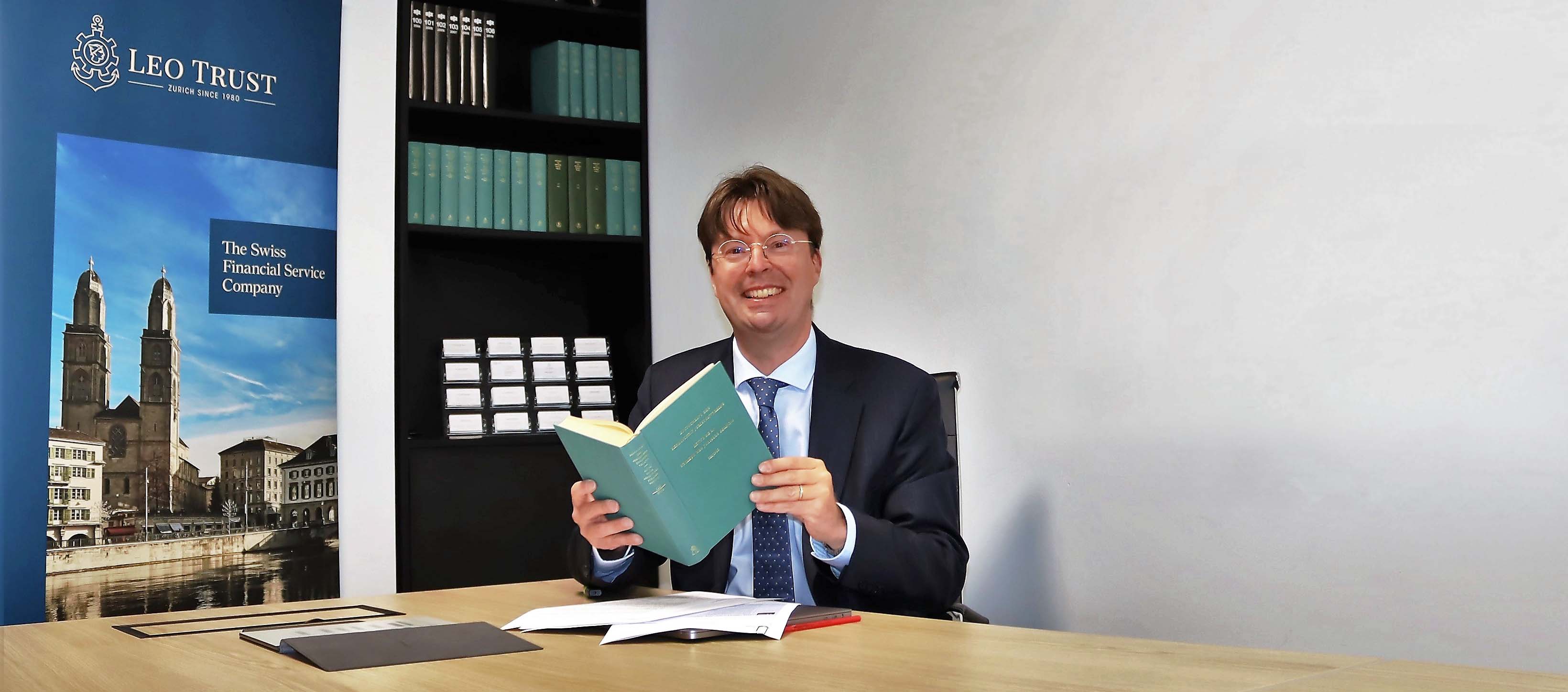 Man in a suit reading a book at a desk in front of bookshelves