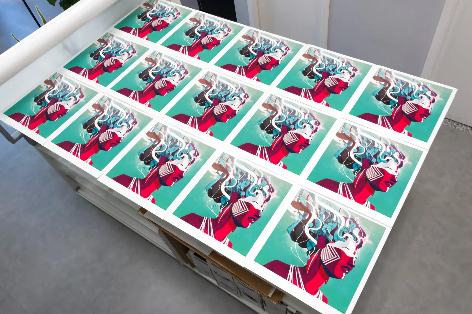 Multiple prints of a stylized, futuristic profile of a person with red skin and flowing white hair on a turquoise background arranged on a table.