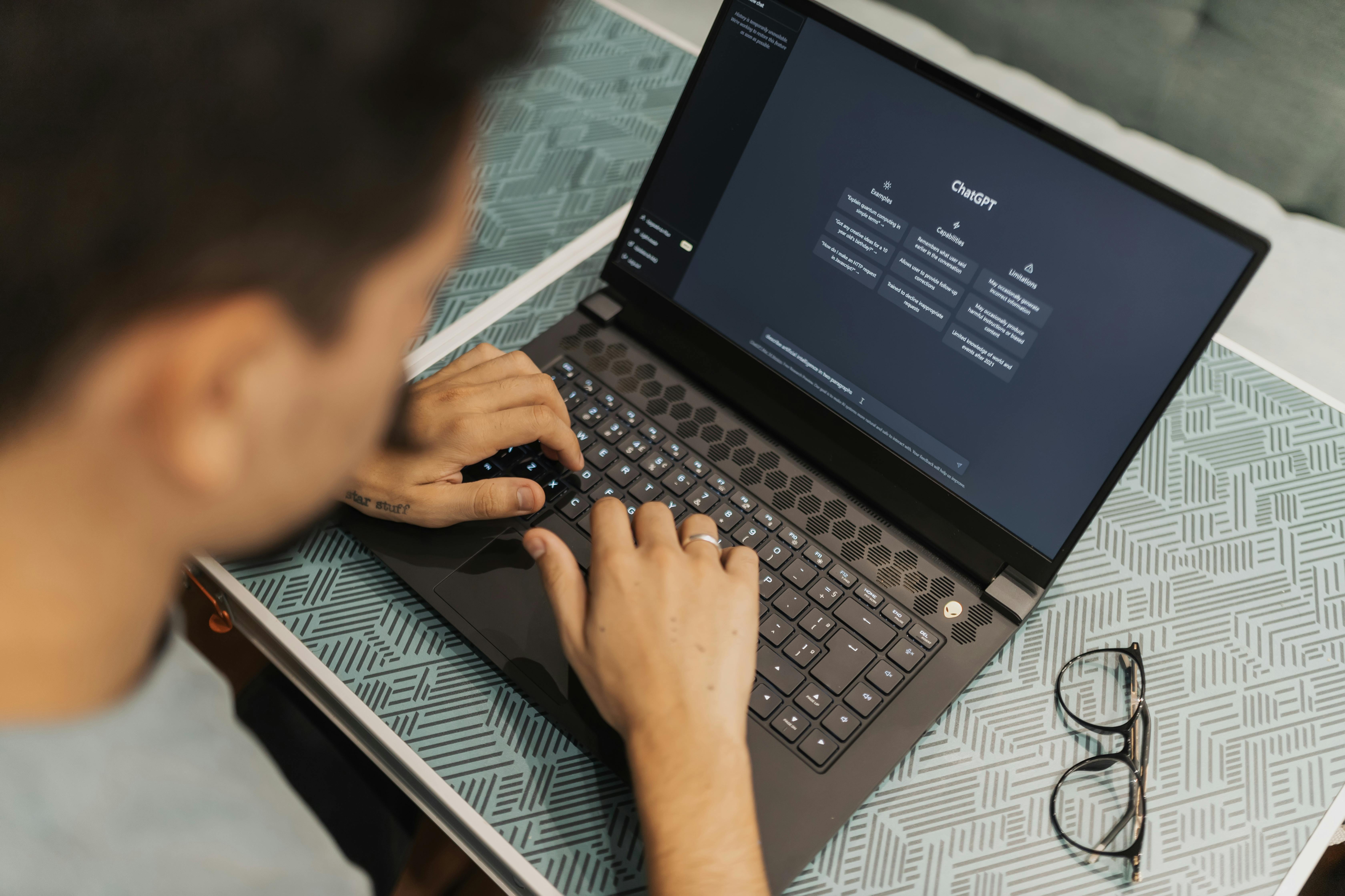 Person typing on a laptop with ChatGPT open on the screen, glasses placed on the table nearby