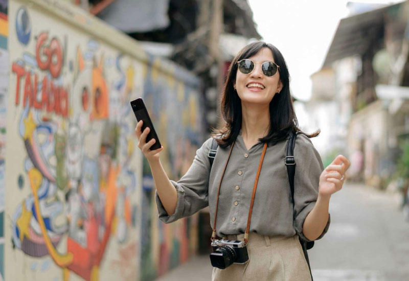 A female travel influencer wearing a pair of sunglasses and holding a smartphone on her right hand, enjoying a stroll in the middle of an art street.