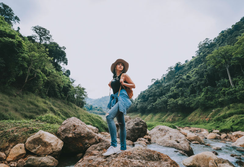 A female adventure traveler wearing a black sleeveless top, a pair of blue jeans and a hat standing on top of a huge stone at the side of a stream.