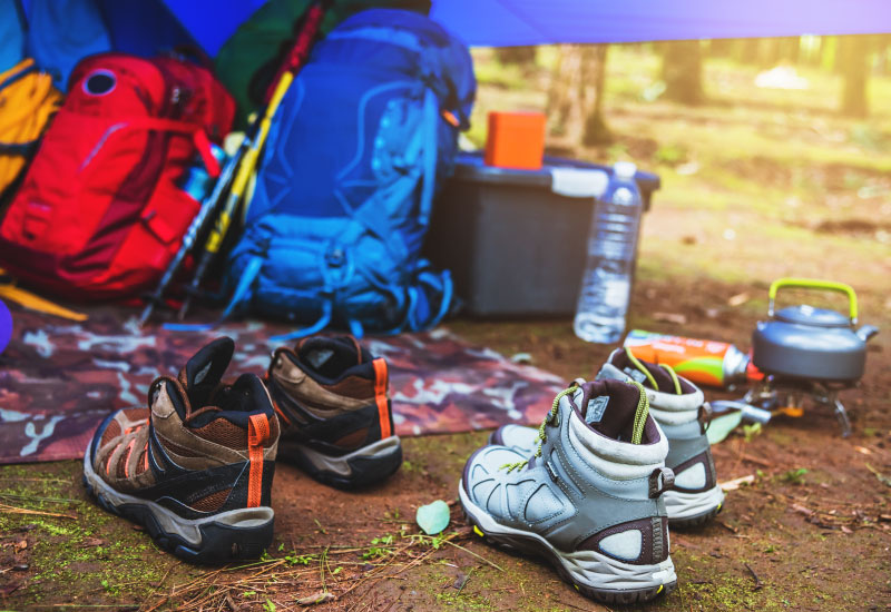 various camping gear including backpacks, water bottle, kettle, hiking shoes, hiking sticks, and a mat under a tent. 