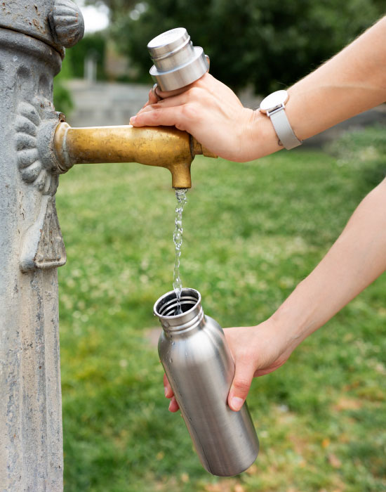 A woman filling up a canned bottle from a water fountain at the park.