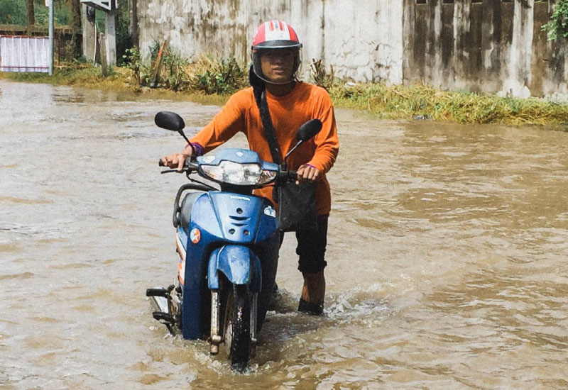 A man in orange tshirt is pushing his stalled motorbike through shin-high flood water.