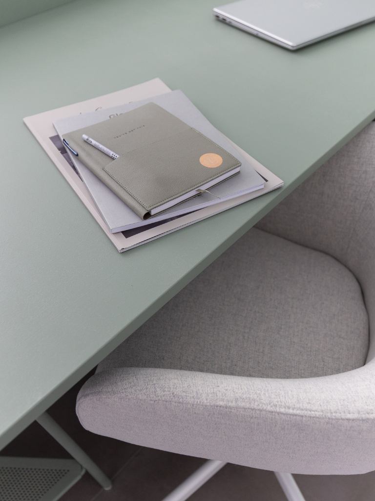 Notizbuch, Notizzettel und Stifte auf einer mintgrünen Tischplatte, davor steht ein gemütlicher Schreibtischsessel mit Stoffbezug in weiß-beige.