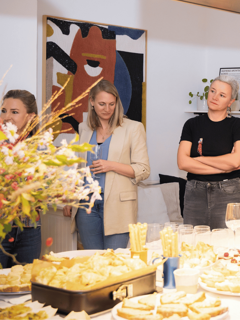 Drei Powerfrauen stehen hinter einem Buffet und hören einem Vortrag zu.