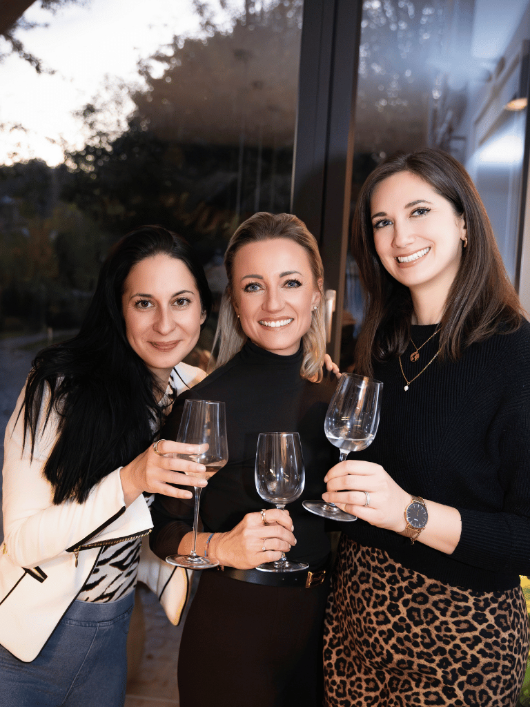Drei Frauen mit Gläsern in der Hand lächeln in die Kamera.